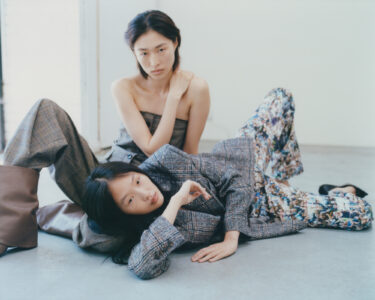 Two individuals are posed in a minimalist setting. One sits behind the other, wearing a strapless outfit, displaying a composed expression. The other is reclined, wearing a plaid jacket and floral pants, gazing thoughtfully. The scene conveys a serene yet stylish atmosphere, with a focus on fashion and intimate connection between the subjects against a simple backdrop.