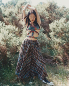 A woman stands in a natural setting with dense shrubbery, basking in sunlight. She wears a colorful plaid top with multiple straps, coordinated with a full, flowing skirt. Her arms are elegantly raised above her head, and her long dark hair cascades down, swaying in the gentle breeze. She gazes intently, exuding a serene confidence.