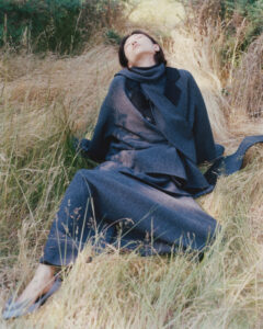 A person is reclining gracefully amidst tall, golden grass. They are cloaked in a long, dark overcoat, and their face is gently tilted upwards, suggesting a serene moment. The natural surroundings create a peaceful atmosphere, with sunlight softly filtering through the foliage and casting gentle shadows. The scene evokes tranquility and a connection with nature.