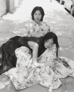 Two individuals are captured in a serene outdoor setting, where dappled sunlight creates patterned shadows. One person sits upright, wearing a floral outfit, exuding calmness. The other leans gently, resting with eyes closed, draped in flowing flower-patterned fabric that cascades around them. The atmosphere is tranquil and reflective, with a peaceful expression and elegant composition.