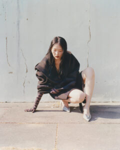 A person is positioned low to the ground, dressed in a dark, draped outfit. Their long, dark hair complements the attire, and they wear formal shoes. The ground consists of large paving stones, and a textured wall with visible cracks serves as a backdrop. Light and shadow add depth to the scene, creating an intriguing composition.
