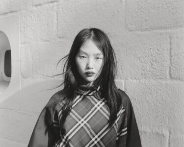 A young woman with long, dark hair gazes directly ahead, her expression serene yet strong. She wears a patterned garment featuring bold diagonal stripes. The setting appears minimalist, with textured, light-colored walls casting subtle shadows. Natural light creates contrasts, highlighting her features and the fabric's details, creating a harmonious balance of intensity and tranquility.