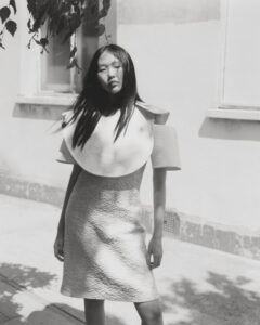 A person stands outdoors wearing a textured dress with an exaggerated collar and shoulder design. The scene is sunlit, casting soft shadows on the ground. The individual has long hair and maintains a relaxed posture. Nearby, leaves dangle from a branch, adding a natural element to the setting. The background shows a wall with a window.