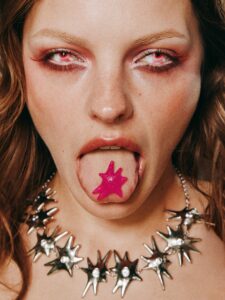 A tightly framed portrait centres on a person staring directly ahead with an intense, almost otherworldly expression. Dramatic eye makeup and vivid red accents draw attention to the gaze, while a bright star-shaped object rests on the extended tongue. A sculptural necklace of metallic spikes encircles the neck, adding edge and tension. The composition feels provocative, surreal, rebellious, and unapologetically bold.