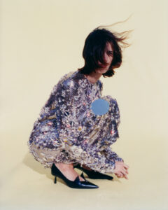 A person with shoulder-length dark hair and a windblown style crouches against a light background. They wear a shimmering, sequined outfit full of intricate patterns and textures, reflecting light from various angles. Their feet are adorned with sleek black heels. The overall look conveys a sense of elegance and contemporary fashion, with a focus on bold details.