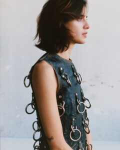 A person with shoulder-length dark hair is shown in profile, sporting a sleeveless, dark garment adorned with large metal rings. The garment appears to be made of a textured material. A small tattoo can be seen on their arm. The background is a muted, faded gray, suggesting an industrial or minimalist setting. The overall tone is edgy and artistic.