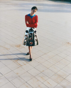 A person stands on tiled ground, casting a long shadow. They're dressed in a red top with blue accents and a striking metallic skirt featuring circular cutouts. The skirt's reflective surface catches light, contrasting with the plain background. Their pose is relaxed yet thoughtful, with arms crossed and feet in black heels, emanating a modern, avant-garde vibe.