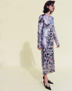 A person stands against a light yellow background, wearing a long, shimmering dress adorned with sequins in various floral patterns. The outfit, with long sleeves and a modest neckline, reflects light, creating an elegant, sparkling effect. The individual has short, dark hair and wears black pointed heels. A subtle shadow is cast on the floor.