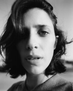 A monochrome portrait captures a person gazing directly with a contemplative expression. The close-up highlights their short, windswept hair and natural skin texture. Shadows add depth around their eyes and mouth, enhancing the serious tone. The background is softly blurred, keeping the focus on the intense expression and delicate play of light and shadow across their face.
