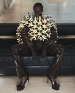 A person sits confidently on a dark leather sofa, wearing an extravagant outfit featuring a striking, colorful disc arrangement covering the upper torso. The ensemble is paired with dark leather pants, gloves, and heeled shoes. The background has a minimalist, muted tone, providing contrast to the vibrant outfit. The composition feels bold and avant-garde.