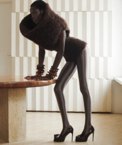 A person stands in a striking pose, wearing a voluminous, textured garment that resembles a large collar. They balance on glossy, high-heeled shoes, while leaning on a wooden surface. The background features a geometric, white, wavy pattern. Bold bracelets adorn both wrists, adding to the overall fashion-forward, avant-garde aesthetic of the scene.