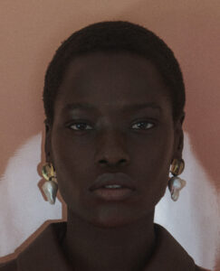 A person with short hair and a neutral expression gazes forward. Their skin has a rich, deep tone. They're wearing distinctive earrings with irregular, organic shapes and a round component. The attire is a dark collared garment. The background is a smooth, muted color, enhancing the focus on the subject's face and jewelry.