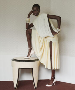 A person poses confidently, dressed in a unique, flowing yellow outfit with fringe details. They are standing with one leg on a round, cream-colored table, accentuating their elegance. The ensemble is completed with multiple bracelets and white high heels. The background features a geometric pattern on the wall, adding a modern, stylish touch to the scene.