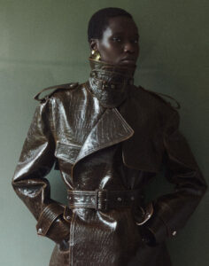 The scene features a person in a high-fashion, textured, dark outerwear piece with distinctive buckles and a high collar. The attire gives off a luxurious, edgy vibe, complemented by large, bold earrings. The backdrop is a muted, soft green, offering a sophisticated contrast to the ensemble. The mood is contemplative and stylish, showcasing modern elegance.