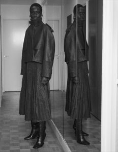 A person stands in a hallway with wooden flooring, wearing a textured, long skirt, high leather boots, and a structured leather jacket. The person faces forward with a serious expression, and their reflection is visible in mirrored panels on the right. The surrounding walls and doors are simple, creating a minimalist, monochromatic scene.