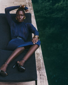 A seated individual reclines on a dark chaise lounge, set against a backdrop of textured stone and lush greenery. Dressed in a fitted blue outfit with high heels, their pose is relaxed yet confident. Accessories include gold earrings and bracelets that add an elegant touch, while an arm is placed behind their head, contributing to the poised demeanor.