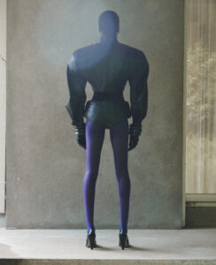 A person stands facing a textured wall, dressed in an avant-garde outfit. The ensemble includes a structured jacket with exaggerated shoulders, dark purple tights, and high-heeled boots. The setting appears to be an understated, architectural space, with muted tones and soft lighting enhancing the dramatic attire. The person's back is straight, contributing to the striking silhouette.