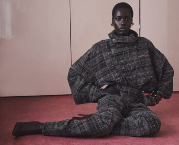 A person is stylishly seated on a textured pink floor, dressed in a heavily patterned, long-sleeved outfit. Their attire is complemented by dark gloves and pointed footwear. They wear large, bold earrings. The expression is poised, reflecting confidence. The background features a minimalist, light-colored wall, adding to the sophisticated, artistic vibe.