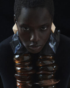 A person with short hair and intense gaze wears striking geometric earrings, combining gold and translucent elements. The dark background highlights a voluminous, layered neckpiece resembling large, amber-toned beads. The overall atmosphere is dramatic and artistic, creating a sense of boldness and sophistication through the use of contrasting textures and deep shadows.