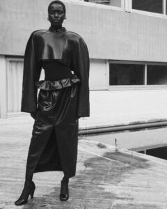 A person stands confidently on a paved surface, wearing a sleek, long black outfit with a dramatic, structured top and a high-slit skirt. The setting has a modern architectural backdrop with large windows. The overall tone is monochromatic and stylish, with a focus on texture and silhouette, creating a striking, high-fashion ambiance.