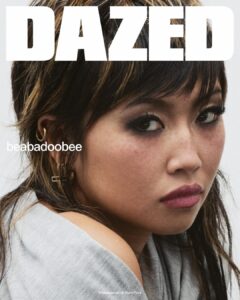 A close-up captures a young woman with soft, tousled hair and layered bangs framing her face. Her gaze turns slightly to the side, expression pensive and introspective. Subtle makeup highlights her eyes and natural skin texture. Multiple small earrings line her ear. Draped in a light fabric, she appears relaxed, while soft lighting enhances a gentle, intimate mood.