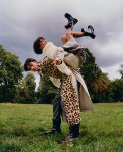 Two individuals are creatively posed in a lush green park under a cloudy sky. One person, wrapped in a leopard print scarf and a long coat, leans forward while the other, dressed in light attire, is playfully balanced upside down on the first person’s back. Black boots on both add a bold contrast to the vibrant setting.