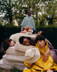 A group of six individuals is gathered outdoors, surrounded by lush green trees. Five of them are lounging on a stack of mattresses, casually draped in colorful and cozy clothing. One person stands behind them, wearing a checkered garment. The scene has a relaxed, whimsical atmosphere, with soft lighting enhancing the natural setting.