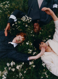 Three individuals are lying in a field of lush green grass interspersed with white wildflowers. Each person wears stylish outerwear in varying shades of dark and light. Their forms create a dynamic composition as they relax, gazing upward. The natural setting contrasts with the structured clothing, creating a harmonious blend of nature and fashion.