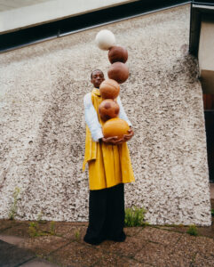 A person stands against a textured wall, balancing a vertical stack of seven balls of different types and colors. They are wearing a bright yellow coat over a white shirt and black pants, holding the bottom ball with both hands. The concrete surface below contrasts with small patches of green plants nearby.