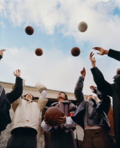 A group of people stand outdoors, reaching upward as several balls are mid-air against a backdrop of a cloudy sky. They are wearing various jackets and coats, looking skyward with a sense of anticipation. The scene conveys energy and movement, capturing a moment of playful activity and communal interaction. Buildings are visible in the background.