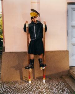 A person stands on wooden stilts, leaning against a pastel pink wall. They wear a large, angular yellow hat and a dark, zippered jacket. Their outfit includes a knee-length, voluminous skirt paired with shiny black boots. The ground is cobbled, and a white door is partially visible to the side, contributing to a whimsical, avant-garde scene.