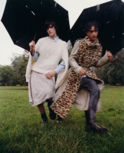 Two individuals walk on grass, each holding a large black umbrella. They are dressed in distinctive outfits: one wears a textured white sweater with a skirt, the other dons a leopard-patterned garment with a long coat. The scene is set outdoors on a cloudy day, suggesting rain, with blurred motion conveying a sense of urgency or movement.