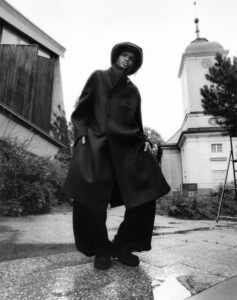 A person stands confidently on a textured pathway, dressed in a long, dark coat and wide-legged pants. They wear a matching hat, tilting slightly in a dramatic pose. Behind, a tall structure with a clock and a spire adds architectural interest. Bushes and a modern building frame the scene, creating a striking contrast between fashion and environment.