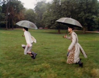 Two individuals sprint across a lush, green field, each holding an umbrella. One wears a long coat with a leopard print detail, while the other sports a white sweater and plaid trousers. The scene is set in a park with tall trees in the background, evoking a whimsical and dynamic atmosphere as they move briskly through the rain.