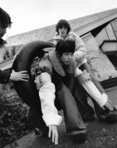 Three individuals are positioned outdoors near an angular building. The central person is wearing an unusual outfit with an oversized black ring around their waist and appears to be crouching. The other two stand by, interacting with the central figure. The scene conveys a sense of playful experimentation and fashion exploration in a grayscale setting.