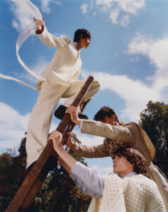 Three people are engaged around a wooden ladder. One, wearing a white outfit, stands atop, arms outstretched with fabric in the breeze. The other two, in patterned attire, brace the ladder amidst a clear blue sky and leafy trees. Motion and balance are central, with dynamic postures conveying support and collaboration under the sunny, cloud-speckled sky.