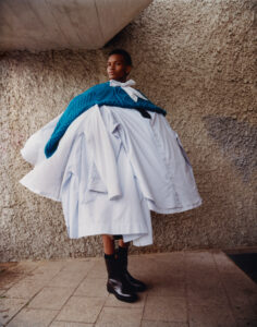 A person stands against a textured concrete wall, wearing a striking, oversized ensemble composed of numerous white and striped shirts layered together. A vibrant blue knit drapes over the shoulders, tied with a white ribbon. Black boots complete the look, adding contrast to the pastel, voluminous garments. The setting is urban, with angular architectural elements.