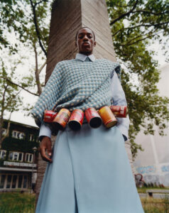 A fashion-forward individual stands beneath a cloudy sky, embodying bold style with a checkered blue sweater layered over a light blue shirt. A unique accessory involves several colorful cans attached creatively around the waist, adding vibrancy. The backdrop features urban elements, trees, and hints of graffiti, infusing an artistic, edgy atmosphere into the scene.