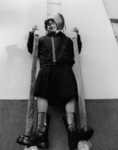 A person with curly hair smiles warmly while standing on tall stilts. Wearing a textured jacket, knee-length shorts, and chunky black boots, they hold the stilts with both hands. The background features a plain, light-colored wall with a small section of a doorway visible. The scene conveys a playful and adventurous atmosphere with an unusual perspective.