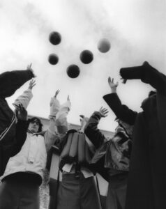 Five people reach upwards towards several floating balls in a monochrome setting. The scene captures a dynamic moment, with each person dressed warmly in various jackets and coats. The angle provides an upward perspective, emphasizing the contrast between the dark clothing and the lighter sky. The composition conveys a playful, energetic atmosphere within an urban environment.