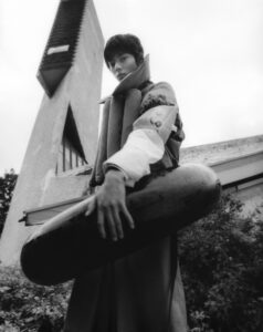 A person stands with a confident expression, wearing a layered outfit with distinct textures. They hold an elongated, cylindrical object. In the background, an angular, modern architectural structure extends upward against a cloudy sky. The scene has a monochromatic palette, blending natural elements with urban design, creating an intriguing juxtaposition and a dramatic, stylish ambiance.
