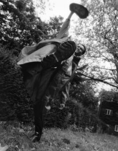 A person executes a high kick in an outdoor setting. The perspective emphasizes the sole of their boot, showcasing the intricate tread pattern. They are wearing a long coat, and the scene features abundant greenery, including trees with dense foliage and shrubbery. A building partially covered in ivy can be seen in the background, suggesting a natural environment.