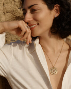A person with short, curly dark hair smiles while resting their chin on their hand. They wear a white blouse, a gold pendant necklace, and earrings. The background reveals a textured, earthy surface. Sunlight gently illuminates their face, enhancing their relaxed, joyful expression. Their hand is adorned with rings, adding a touch of elegance to the serene scene.