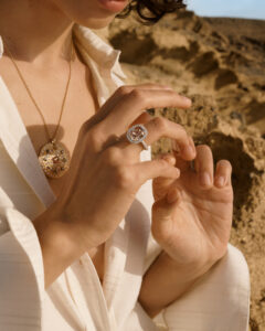 A person with short hair wears a white, textured blouse, open at the chest. They showcase a hand adorned with a large, ornate ring featuring a central gem encircled by smaller stones. A necklace with a large pendant complements the jewelry. The person is positioned against a backdrop of rocky terrain under a clear blue sky.