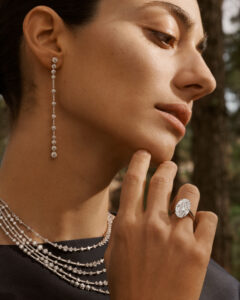 A woman gazes thoughtfully, adorned in elegant jewelry. Her long, delicate drop earring features glistening diamonds, perfectly complementing the multi-layered diamond necklace. She gently touches her chin, showcasing a striking, large diamond ring on her finger. The scene exudes sophistication and grace, with the background hinting at a natural setting, enhancing her timeless beauty.
