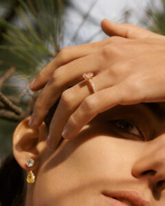 A person is partially covered by their own hand, adorned with a delicate diamond ring featuring a pink gem on a golden band. The light softly highlights two earrings; one with a clear stone, and another with a pear-shaped yellow gem. A background of blurred green foliage adds a natural contrast, creating an elegant and serene scene.
