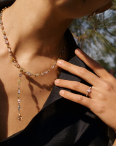 A person is adorned with a delicate necklace featuring various colored gemstones. Their hand gently rests on their chest, showcasing a sparkling ring with a light pink central gem. The background hints at an outdoor setting with soft, natural lighting, casting a warm glow on the skin and jewelry, enhancing the luxurious feel.