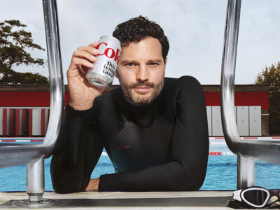A man with curly hair leans against a pool ladder wearing a black wetsuit. He holds a silver can, subtly angled towards the viewer. Behind him, the clear blue water contrasts with red and white pool barriers. Trees and a building with horizontal panels frame the scene. Nearby, white goggles rest on the pool's edge.