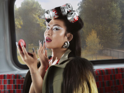 A stylish woman with elaborate makeup and striking silver nails holds a compact mirror. Her hair is creatively styled using soda cans as curlers. She wears bold, oversized earrings and a fur-trimmed coat. The setting is a modern, red-patterned seat near a large window, with a blurred view of lush greenery in the background.