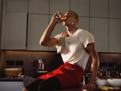 A person is sitting on a kitchen counter, drinking from a can. They wear a white shirt and a red apron, with a towel draped over one shoulder. The background shows cabinets and a countertop with kitchen items like bowls, a mixer, and utensils. The lighting casts gentle shadows, creating a warm, cozy atmosphere.
